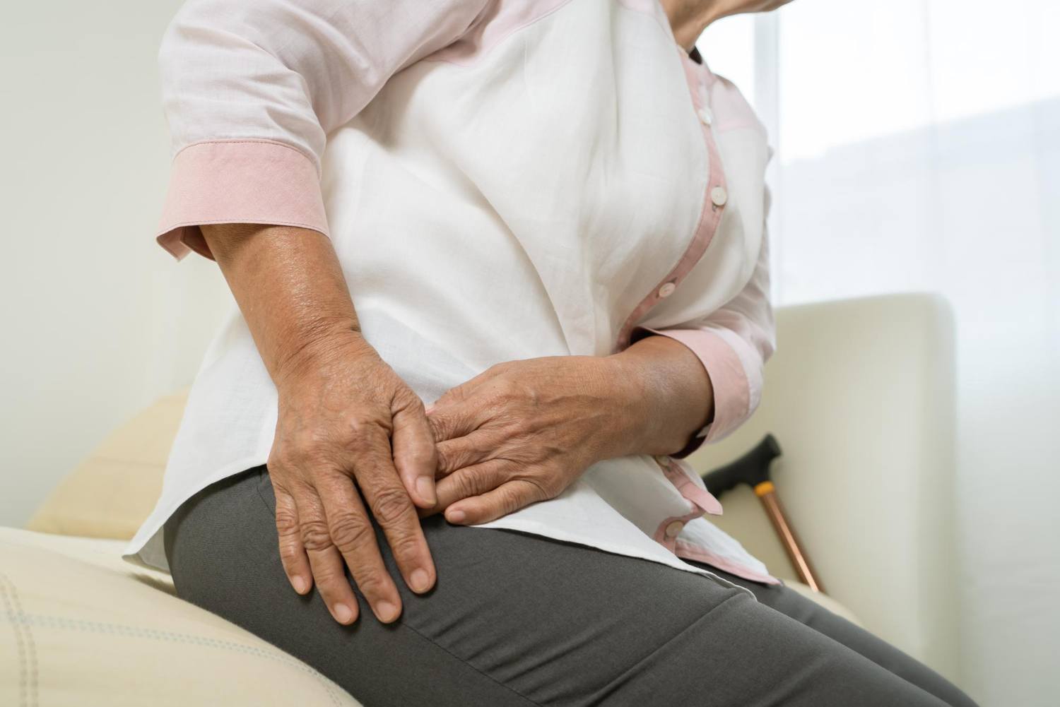 An image of a woman who is sitting with both hands near her right hip. A cane is leaning near her other side at the back of the image. Hip replacement is a medical treatment that may be recommended if non-surgical support is not adequate for an individual.